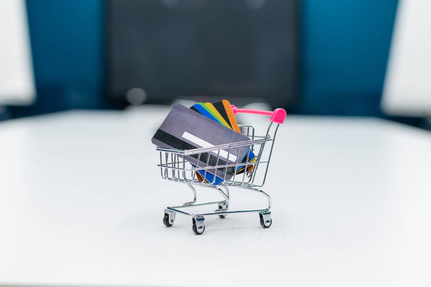 A mini shopping trolley full of credit cards stands on a white table. No people. Close-up of a cart with several different bank debit cards. - Foto, Imagem