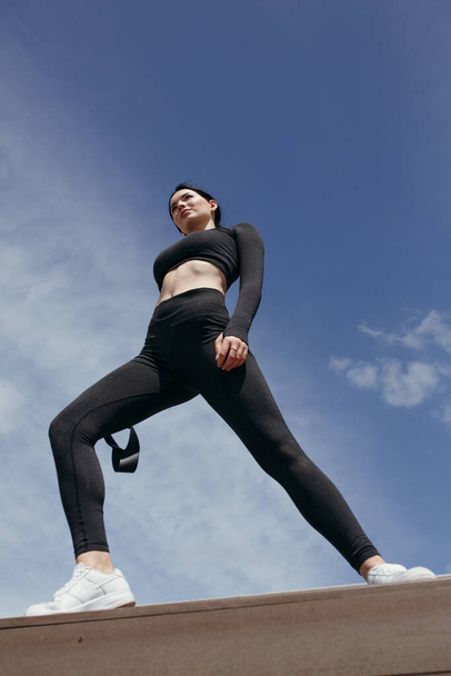 Bottom view of a pilates trainer in a black tight-fitting tracksuit doing exercises with a black elastic expander tape - Foto, afbeelding