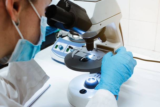 青い手袋とマスクで医療用顕微鏡を通してサンプルを見ている科学者。ウイルス病パンデミック。風景写真. - 写真・画像
