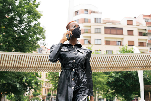 Красивая модель черной девушки в маске из-за пандемии коронавируса ковид 19 разговаривает по мобильному телефону, заботясь о своем бизнесе на улице - Фото, изображение