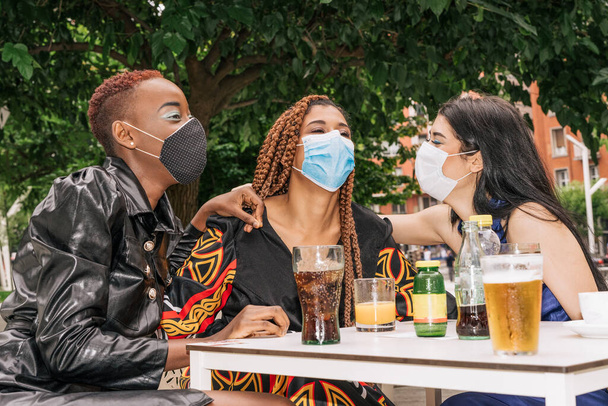 três multirraciais, negros, misturados raze e amigos caucasianos em um terraço bar beber uma bebida enquanto se diverte com máscaras faciais devido ao coronavírus covid 19 pandemia - Foto, Imagem