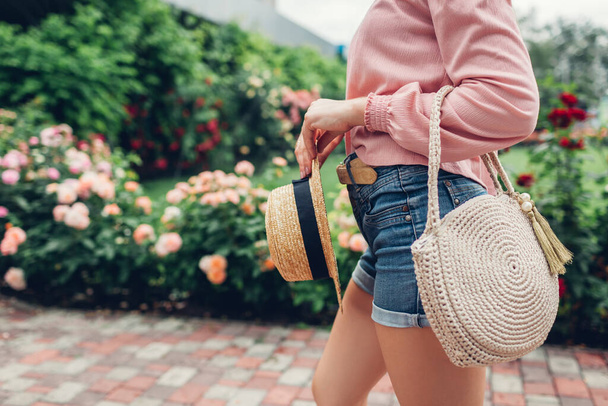 Stílusos női kézitáska és szalmakalap. Fiatal nő visel ruhát gazdaság gyönyörű nyári kiegészítők a kertben szabadban a rózsák virágok a parkban. Városi divat - Fotó, kép