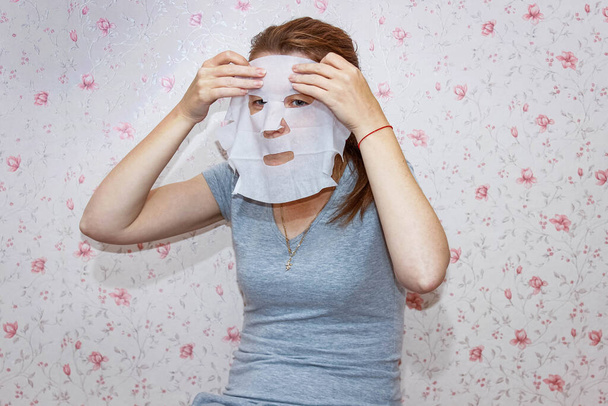 A young woman at home puts a cloth cosmetic mask on her face. Skin care. - Fotoğraf, Görsel