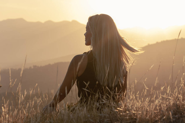 Egy lány ül a fűben egy dombtetőn csukott szemmel, és meditál a hegycsúcsok és a naplemente hátterében, szőke haja repül a szélben.. - Fotó, kép