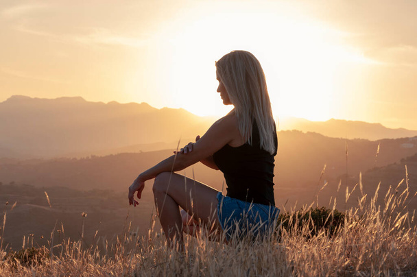 Vue latérale d'une fille blonde en T-shirt, short et baskets accroupi entre les oreilles d'herbe sur fond de soleil couchant et de sommets montagneux. - Photo, image