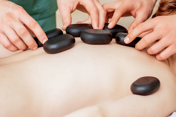 Männchen vier Hände legen große neue ovale schwarze heiße Steine für Rückenmassage auf dem weiblichen Rücken im Wellness-Salon - Foto, Bild