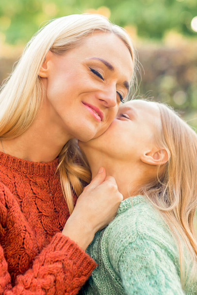 Cute daughter kisses her young mother while sitting at the park - Photo, Image