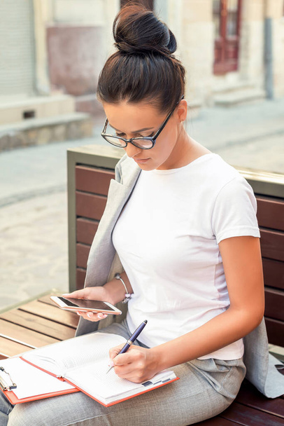 Piękna młoda kobieta robi notatki w notatniku, siedząc na ławce na ulicy. Koncepcja stylu życia - Zdjęcie, obraz