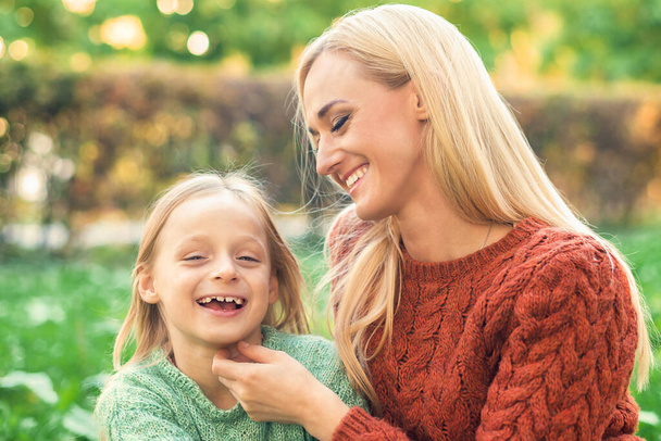 Όμορφη νεαρή καυκάσια μητέρα και η μικρή κόρη χαμογελούν μαζί στο πράσινο γρασίδι στο Φθινοπωρινό Πάρκο - Φωτογραφία, εικόνα