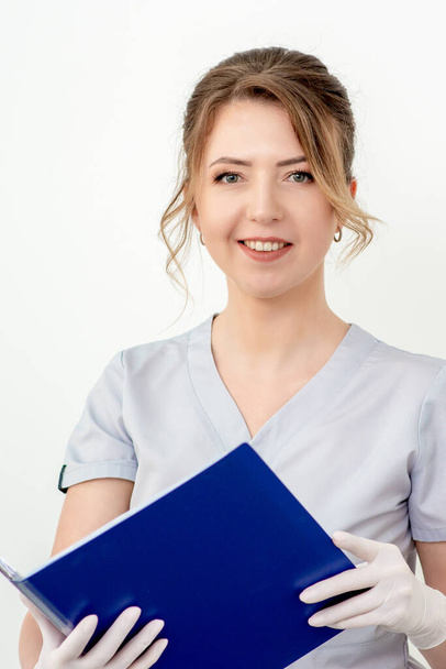 Portrait of beautiful young nurse holding blue folder in gloves looking at camera on white background - Valokuva, kuva