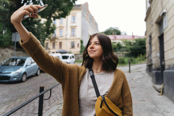 Jeune femme en tenue décontractée à la mode prenant selfie tout en marchant dans la rue de la ville. Élégant hipster fille tenant téléphone et bavarder en ligne. Communication sociale - Photo, image