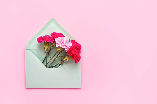 ピンク色のパステル地に緑色の封筒にピンクのカーネーションの花を咲かせます。誕生日、母の日、独身、結婚式のコンセプトのフラットレイアウト。スペースをコピーし、上のビュー。グリーティングカード - 写真・画像