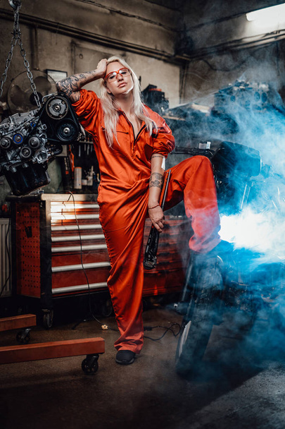 Blond female mechanic with tattooed hands in orange overalls stands in garage or workshop - Fotó, kép