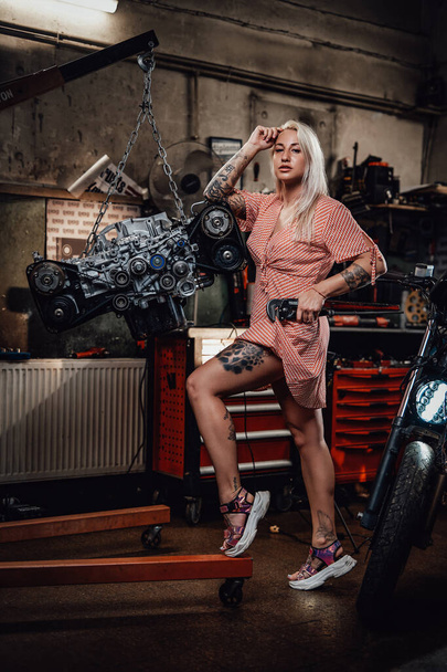 Beautiful blonde girl with tattooed body wearing pink dress posing in garage or workshop - Photo, Image