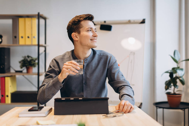 Boldog üzletember, aki laptoppal dolgozik az irodájában. Fiatal mosolygós férfi ül a munkahelyén notebook számítógéppel és kezében pohár vízzel - Fotó, kép