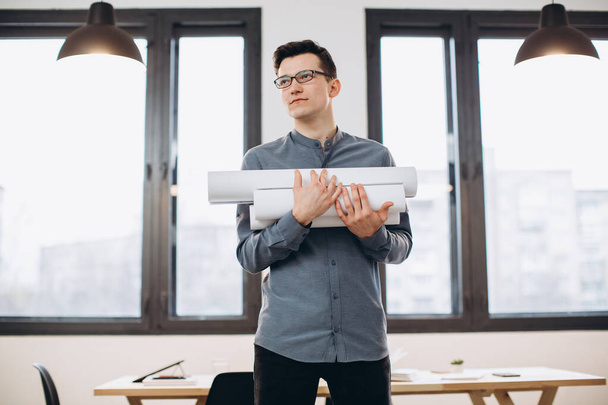 Attractive young man architect in glasses standing isolated over loft office background, carrying drawings, holding house scheme - Valokuva, kuva