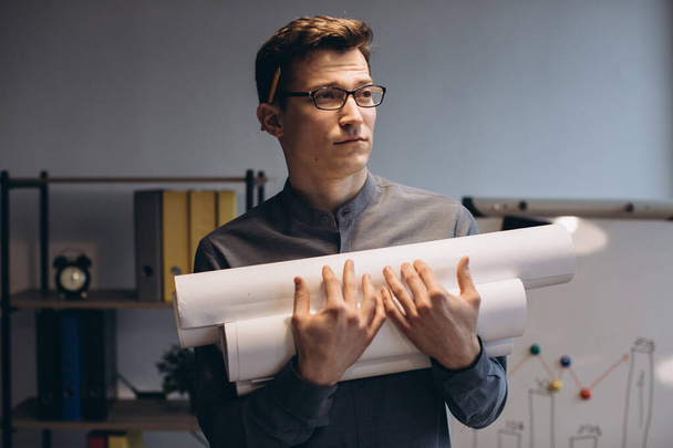Attraktiver junger Mann Architekt mit Brille, der isoliert über Nacht im Büro steht, Zeichnungen trägt, Hausordnung hält - Foto, Bild