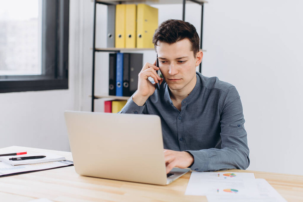 Modern technology and communication concept. Good-looking unshaven young businessman employee working on generic laptop computer and having phone conversation in office - Photo, Image
