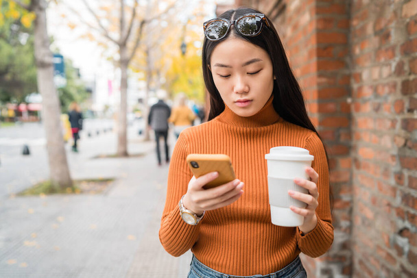 Портрет молодої азіатки, яка користується мобільним телефоном, тримає на вулиці чашку кави. Міське та комунікаційне поняття. - Фото, зображення