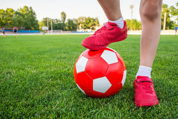 A boy stands on the football field of the stadium and holds his foot on a soccer ball. Children play football on the grass. Training or competition concept. - Photo, Image