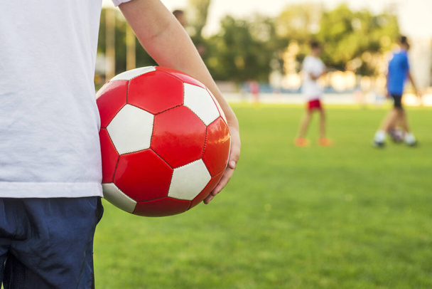 Ένα αγόρι στέκεται στο γήπεδο ποδοσφαίρου του σταδίου και κατέχει μια μπάλα ποδοσφαίρου με φόντο την ομάδα του ή την αντίπαλη ομάδα. Έννοια κατάρτισης ή ανταγωνισμού. - Φωτογραφία, εικόνα
