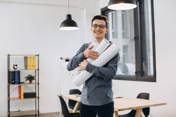 Attractive young man architect in glasses standing isolated over loft office background, carrying drawings, holding house scheme - Foto, imagen