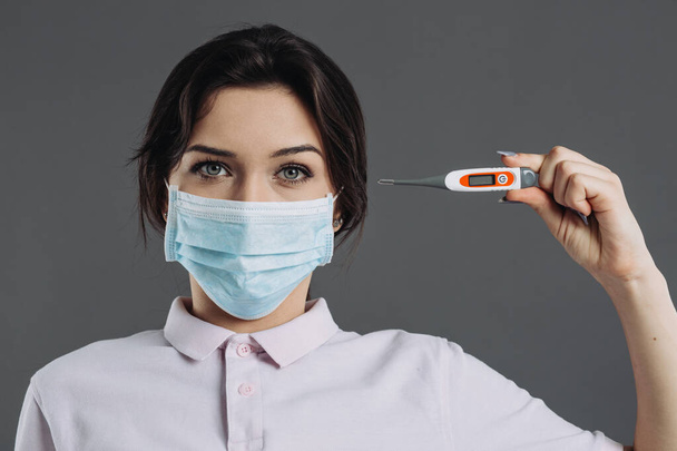 Woman in medical protective mask holds thermometer which shows high temperature. Symptoms of coronavirus - Photo, Image