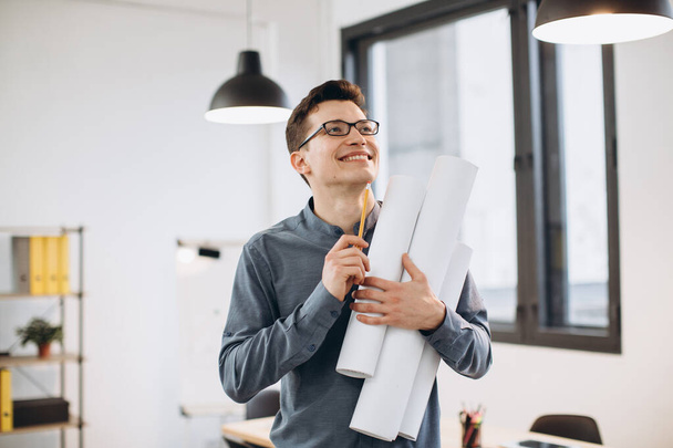 Attractive young man architect in glasses standing isolated over loft office background, carrying drawings, holding house scheme - Foto, afbeelding