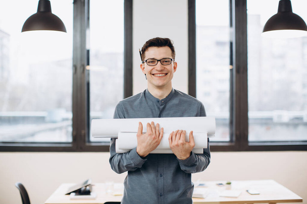 Привабливий молодий чоловік архітектор в окулярах стоїть ізольовано на фоні лофт офісу, несучи креслення, тримаючи схему будинку
 - Фото, зображення