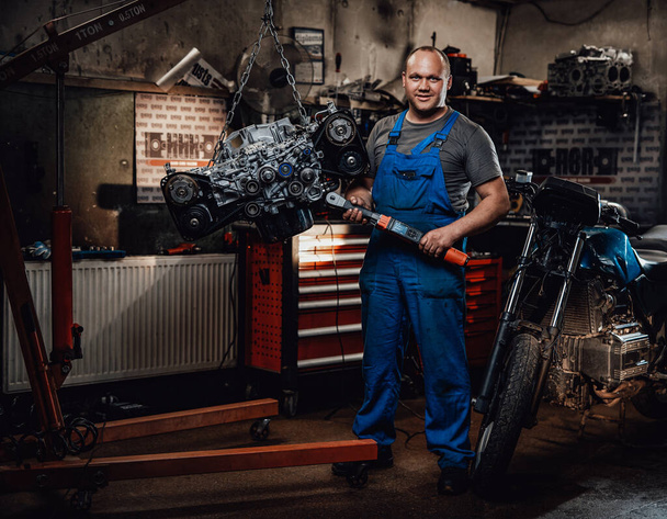 Profesionální mechanik stojí vedle zavěšeného plochého motoru a nahého kola v garáži - Fotografie, Obrázek