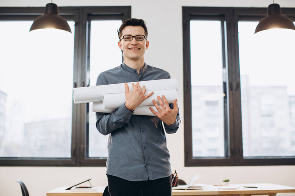 Attraente giovane architetto in occhiali in piedi isolato su sfondo ufficio soppalco, portando disegni, schema di tenuta casa - Foto, immagini
