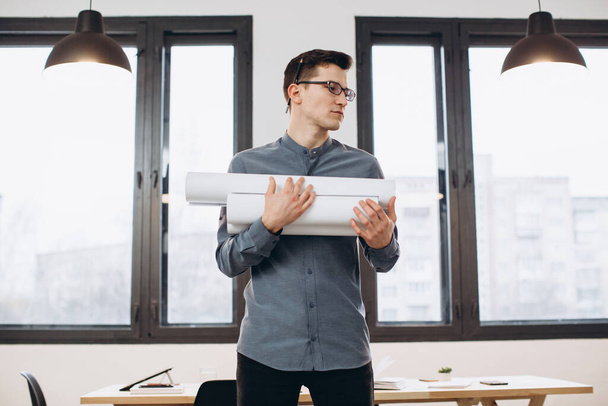 Attraente giovane architetto in occhiali in piedi isolato su sfondo ufficio soppalco, portando disegni, schema di tenuta casa - Foto, immagini
