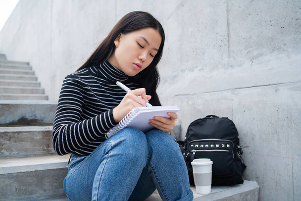 Portrait of young asian woman, writing on notebook while sitting outdoors on concrete stairs. - Foto, Imagen