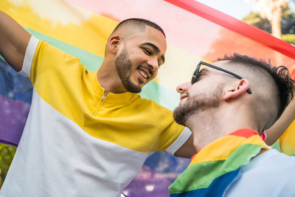 Portrait of young gay couple embracing and showing their love with rainbow flag in the stret. LGBT and love concept - Фото, изображение