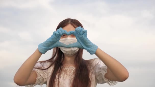 menina em uma máscara protetora e luvas médicas mostra símbolo do coração com as mãos. A jovem adora médicos. pandemia, covid 19. médico feminino mostrando dedos de coração e amor para pacientes. - Filmagem, Vídeo