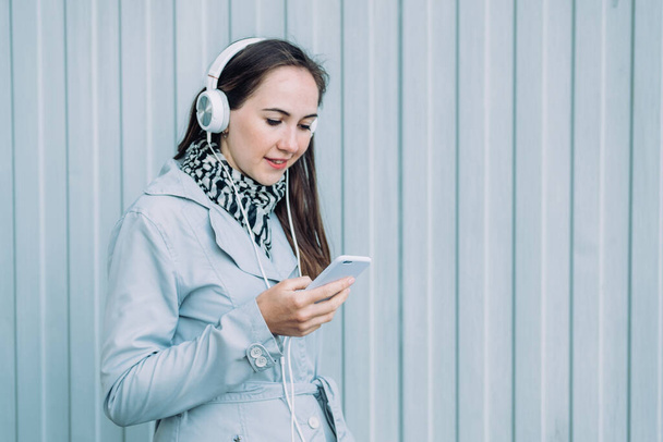 Mulher em fones de ouvido e em um casaco cinza olha para um smartphone
. - Foto, Imagem