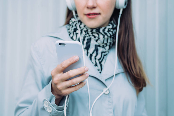 Girl in headphones and in a gray coat listens to an audiobook - Φωτογραφία, εικόνα