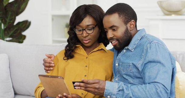 Happy African American couple husband and wife sitting on sofa using tablet and credit card for online shopping at home. - Photo, Image