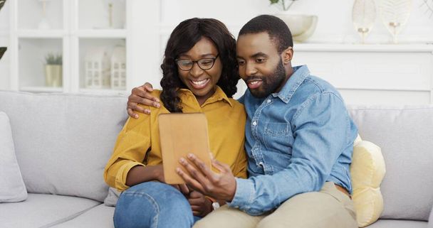 Happy African American couple husband and wife sitting on sofa using tablet at home. - Fotografie, Obrázek