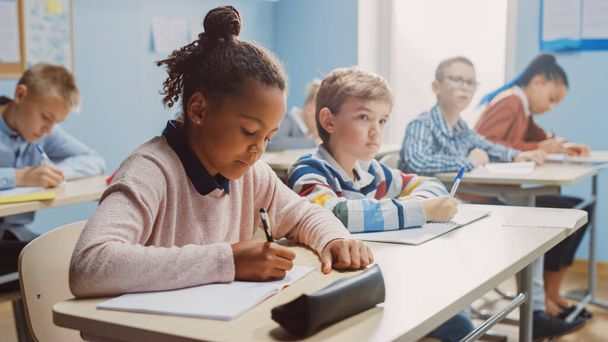 In Elementary School Classroom Brilliant Black Girl Writes in Exercise Notebook, Taking Test and Writing Exam. Junior Classroom with Diverse Group of Children Working Diligently and Learning New Stuff - Foto, imagen