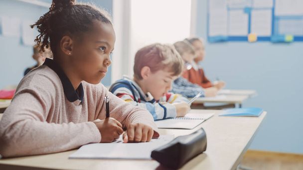 In Elementary School Classroom Brilliant Black Girl Writes in Exercise Notebook, Taking Test. Junior Classroom with Diverse Group of Bright Children Working Diligently and Learning. Side View Portrait - Fotoğraf, Görsel