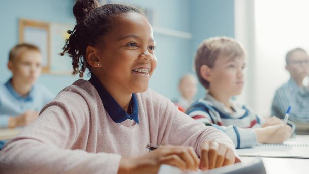 In Elementary School Class: Portrait of a Brilliant Black Girl with Braces Smiles, Writes in Exercise Notebook. Junior Classroom with Diverse Group of Children Learning New Stuff - Photo, Image