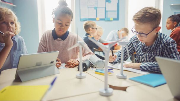 In the Elementary School: Class of Brilliant Young Children Work as a Team Using Tablet Computers to Program Wind Turbines. Classroom with Kids Learning about Eco-Friendly Forms of Renewable Energy - 写真・画像