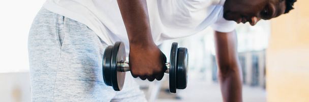 Side view of African American man in sportswear leaning on bench and doing row exercise with dumbbell during training in gym - Фото, зображення