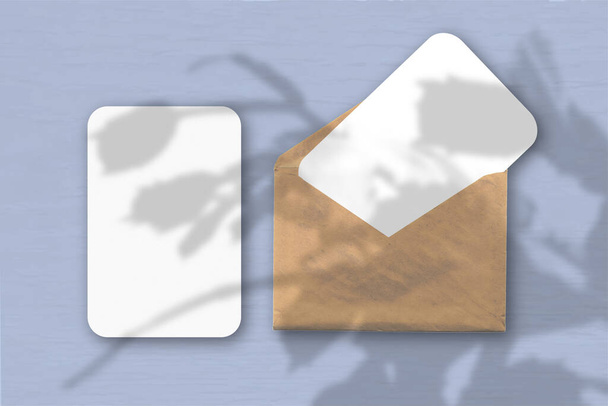Een envelop met twee vellen getextureerd wit papier op een blauwe tafelachtergrond. Mockup met een overlay van plantenschaduwen. Natuurlijk licht werpt schaduwen van een tropische plant. - Foto, afbeelding
