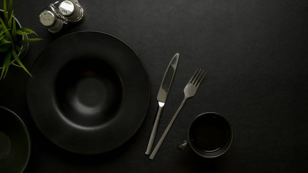 Top view of dark luxury dinning table setting with black ceramic plate, silverware, seasoning bottles, copy space and decoration - 写真・画像
