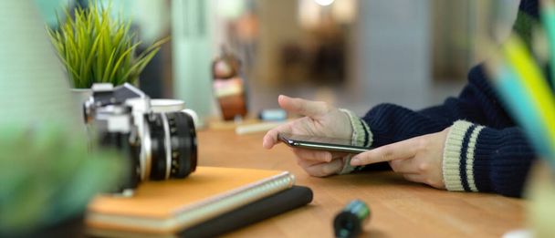 Side view of female hands holding smartphone on wooden home office desk with camera and supplies - Foto, imagen