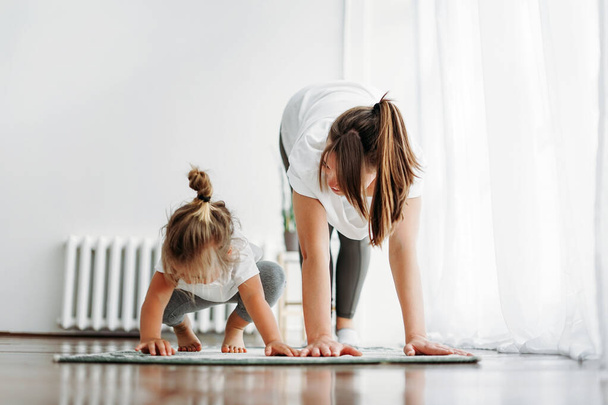 Счастливая мама и маленькая дочь делают утренние упражнения вместе в светлом интерьере дома
 - Фото, изображение