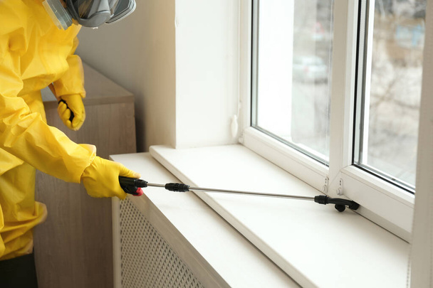 Pest control worker in protective suit spraying pesticide near window indoors, closeup - Photo, image