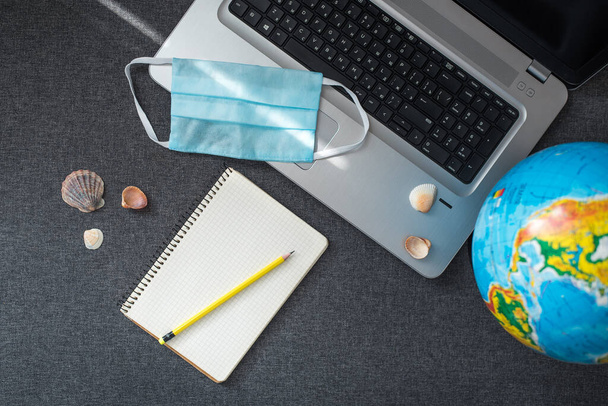 Top view of laptop, notepad, sea shells, globe and  medical mask on the grey background. Concept of working from home in self-isolation, travel business, safe travelling - Photo, Image